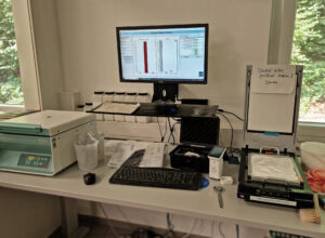 Das OKS Scanner System im Labor von Kröner Stärke. Ein Monitor in der Mitte auf einer Ablage, davor Maus und Tastatur, rechts der Scanner, etwas unaufgeräumt, Pinsel, Staubtücher, Bild aus der Arbeit